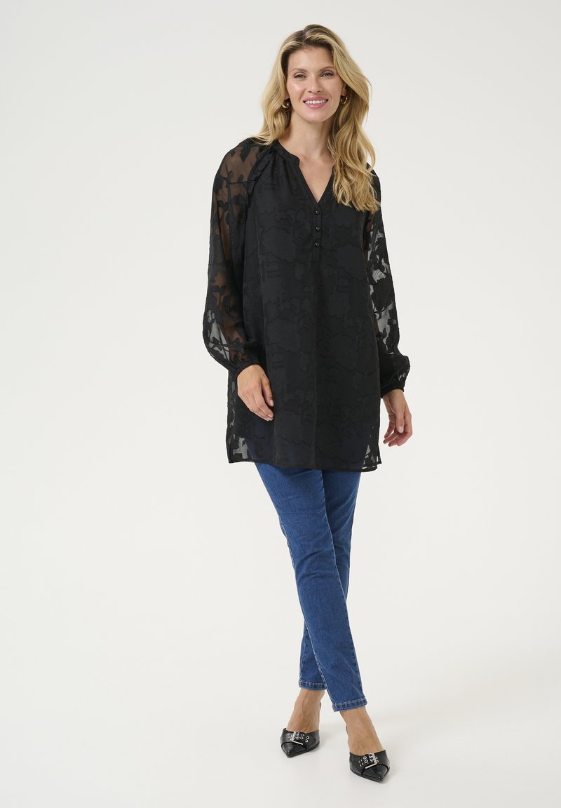 Schwarze transparente Bluse mit floralem Muster, V-Ausschnitt, Knopfdetails und langen Ärmeln. Kombiniert mit blauen Jeans und schwarzen spitzen Schuhen.