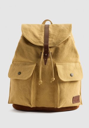 Gelber Canvas-Rucksack mit einer abgerundeten Klappe, braunen Lederelementen und Fronttaschen. Besitzt einen Kordelzugverschluss und ein Logo-Patch an der Unterseite.