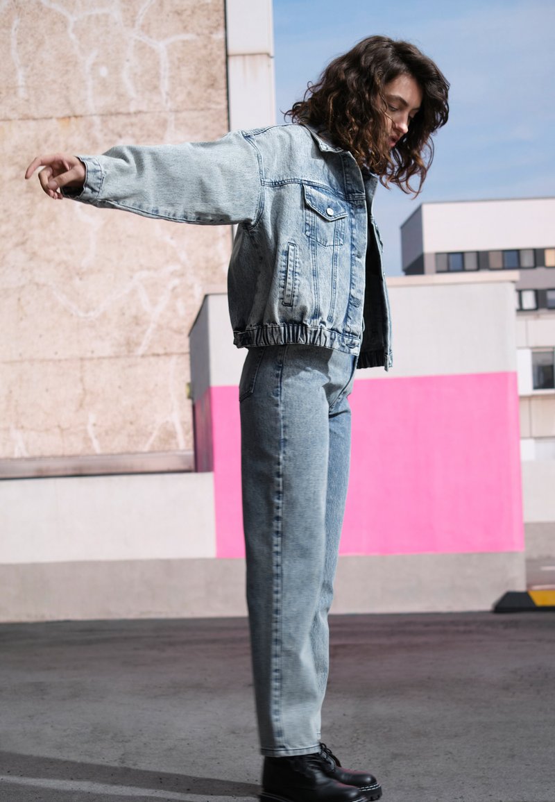 Casaco de ganga e jeans em azul claro, com um design cropped e bainha desfiada. Botas pretas de atacadores completam o visual.