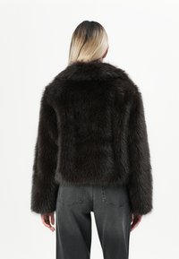 Chaqueta de piel sintética en marrón oscuro con un diseño corto, que presenta un amplio cuello y un material texturizado y esponjoso. Combinada con vaqueros grises.