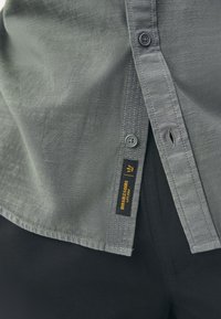 Gray button-up shirt featuring a smooth texture, subtle sheen, and a tag reading "Durable Goods" with a black and yellow design.
