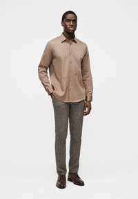 Καφέ πουκάμισο με μακριά μανίκια και γιακά με κουμπιά, συνδυασμένο με υφασμένα γκρι παντελόνια και σκούρα καφέ παπούτσια, στέκεται μπροστά σε λευκό φόντο.