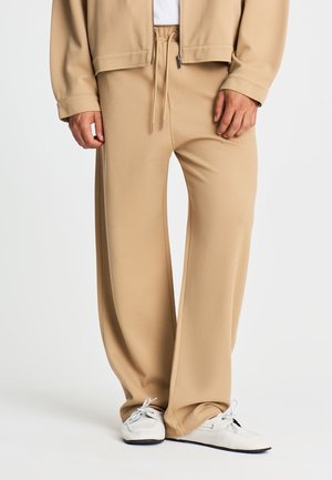 Uomo che indossa pantaloni beige a gamba larga, scarpe bianche e una giacca beige con le mani rilassate lungo i fianchi.