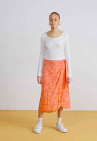 Hvit langermet ribbestrikket topp med rund nakke, kombinert med et lyst oransje blomsterwrapskjørt. Hvite joggesko og ankelstrømper fullfører stilen.