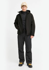 Zwarte waterdichte jas met een capuchon over een zwart thermoshirt, gecombineerd met zwarte broek en geel-zwarte laarzen. De beanie is beige.