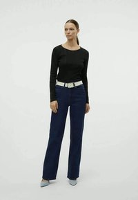 Top preto de manga longa, jeans azuis de cintura alta com um cinto branco, e sapatos apontados azul claro. Modelo está em frente a um fundo branco liso.