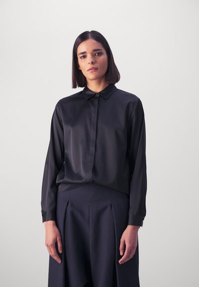 Chemise noire en satin à manches longues avec col, dotée d'un devant à boutons. Portée avec un pantalon bleu marine texturé, mettant en avant un tissu lisse et fluide.