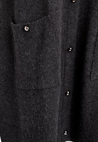 Kepenė tamsiai pilkos spalvos, pagaminta iš storos mezgimo medžiagos, su juodais mygtukais, dviem šoninėm kišenėm su banguotais kraštais ir minkšta tekstūra.