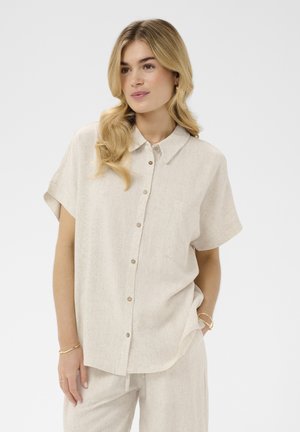 Woman with blonde hair wearing a loose beige short-sleeve button-up shirt and matching pants with minimal gold jewelry.