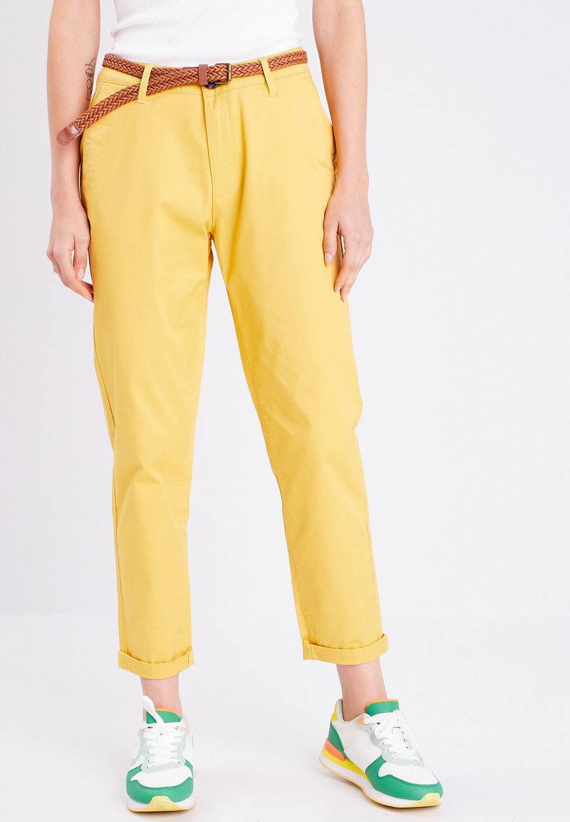 Chino en coton jaunes avec taille haute, ourlets roulés et une ceinture en cuir tressé marron, associés à des baskets vertes et blanches.