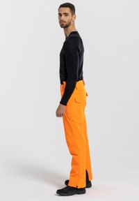 Oranje cargobroek met zijzakken en verstelbare banden, gecombineerd met een aansluitend zwart longsleeve shirt en zwarte schoenen.