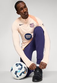 Athlete wearing a pale peach long-sleeve shirt with a Barcelona logo and dark purple pants, tying black shoes beside a multicolored soccer ball.