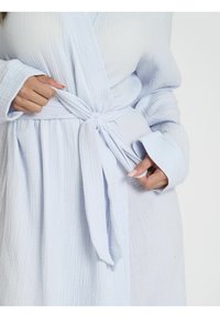 Abito avvolgente azzurro chiaro con texture, cintura a cravatta e maniche larghe. Primo piano del dettaglio della vita e del motivo del tessuto.