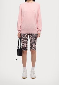 Pull en maille rose à manches longues, associé à un short de cycliste à imprimé léopard noir et rose. Le mannequin tient un petit sac à main noir, portant des baskets blanches.