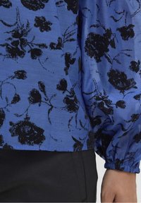 Blusa floreale blu con stampa floreale nera, maniche lunghe a sbuffo e polsini elastici. Tessuto leggero con una sottile texture.