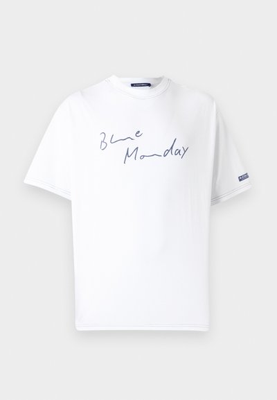 Valge puuvillane T-särk, millel on avar lõike, esitades sinise käsitsi kirjutatud teksti "Blue Monday" ees ja logotägi varrukal.