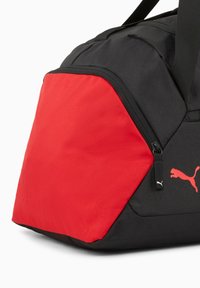 Sort duffel taske med en kontrasterende rød panel, stofmateriale, rektangulær form, har en lynlås og Puma-logo i hvid.