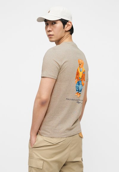 Ανοιχτό καφέ κοντομάνικη μπλούζα με πολύχρωμο σχέδιο αρκούδας στην πλάτη, με το κείμενο «POLO BEAR by RALPH LAUREN» από κάτω.