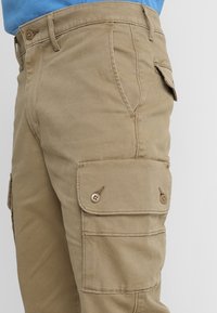 Beige cargo-housut, jotka on valmistettu kestävästä kankaasta, sivutaskuilla, joissa on nappisulku ja suora lahje.