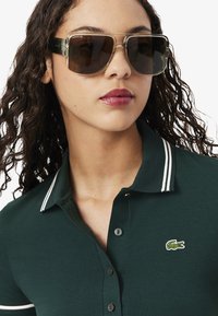 Donkergroene polo met witte gestreepte kraag, voorzien van het Lacoste krokodillenlogo. Model draagt grote, ronde zonnebril met een transparant frame.