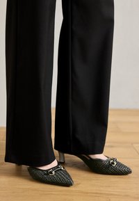 Mules negros de punta afilada con textura trenzada, detalles en tono dorado y un tacón stiletto bajo, combinados con pantalones anchos negros.