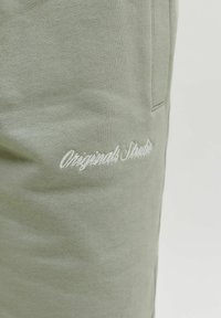 Jasnozielony materiał z białym kursywnym haftem z napisem „Originals Studio” w pobliżu bocznego szwu.