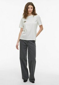 Camiseta de manga corta gris claro con un detalle de labios de leopardo; combinada con pantalones de pierna ancha negros y zapatos negros. Fondo liso.