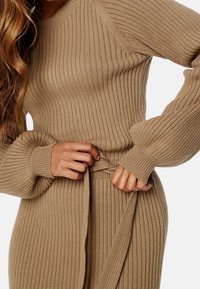 Femme nouant la ceinture d'une robe en maille beige côtelée à manches longues ballon et col montant.