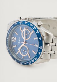 Ασημί ρολόι χρονογράφου από ανοξείδωτο ατσάλι με μπλε καντράν, ροζ χρυσές λεπτομέρειες, περιστρεφόμενη βάση ταχυμέτρου και τρεις υποκαταγραφές. Παράθυρο ημερομηνίας.