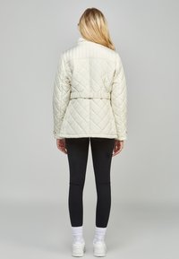 Kvinna som står med ryggen mot betraktaren, iklädd en krämfärgad quiltad jacka med bälte, svarta leggings, vita strumpor och vita sneakers mot en enkel grå bakgrund.