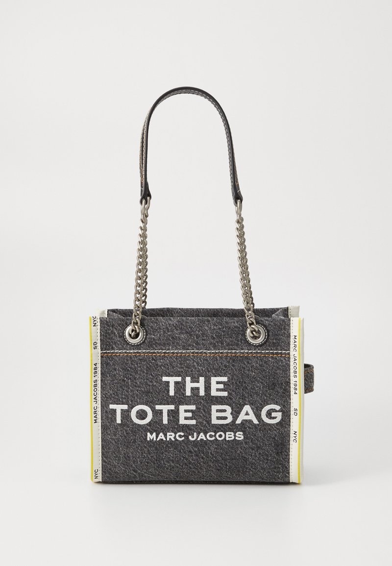 Sac fourre-tout en denim gris avec chaînes en argent et anses en cuir. Présente un texte blanc et des accents jaunes sur les côtés. Forme rectangulaire avec un design robuste.