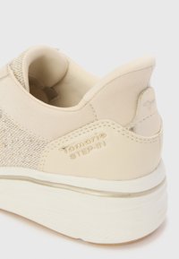 Beige sneaker featuring a textured mesh upper, smooth accents, a subtle metallic strip, and embossed logo on the heel. Rubber sole.
