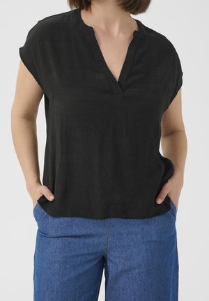 Femme portant un chemisier noir ample à manches courtes avec un col en V et un pantalon en jean bleu, mains dans les poches.