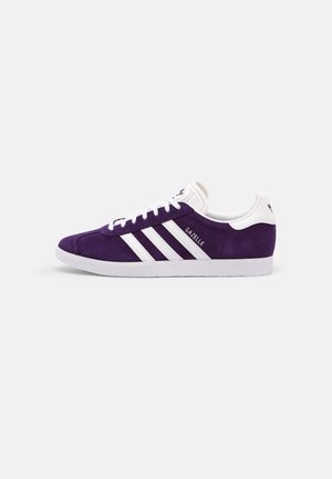 Baskets Adidas Gazelle en daim violet avec des rayures blanches, des lacets blancs et une semelle blanche, vues de côté sur un fond uni.