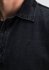 Zwarte denimjas met een gestructureerde kraag, voorzien van een subtiele logo op de borst, met een textuurafwerking en strakke, scherpe lijnen.
