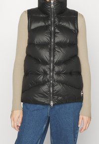 Gilet imbottito nero con colletto alto, chiusura con zip e pannelli cuciti, indossato sopra una camicia beige a maniche lunghe e jeans blu.