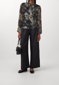 Donna che indossa una blouse floreale trasparente con collo a cravatta, pantaloni neri a gamba larga, ballerine nere, e tiene una piccola borsa nera.