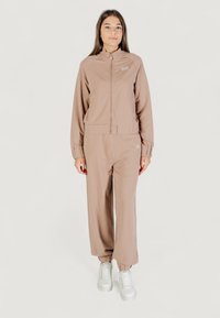 Mujer joven de pie, mirando hacia adelante, vestida con una chaqueta con cremallera de color marrón claro y pantalones a juego, con zapatillas blancas, sobre un fondo blanco.