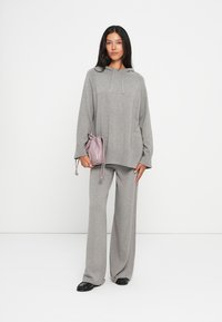 Corta de capuz cinza em malha e calças largas combinando; tecido leve com um corte casual. A modelo segura uma pequena mochila com cordão em rosa claro.