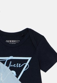 Navy denimsininen lyhythihainen vauvanhaalari, jossa on vaaleansiniset kolmion muotoinen logo ja sana "Guess" kaunokirjoituksella. Napsautettavaa suljinta kauluksessa.