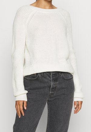 Personne portant un pull en maille blanc à manches longues associé à un jean en denim noir taille haute, debout devant un fond uni.