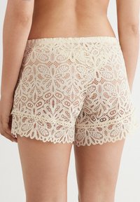 Creme kleurige kantshorts met een bloemenpatroon, een scalloped zoom en een elastische tailleband. De achterkant benadrukt de ingewikkelde details en transparantie.