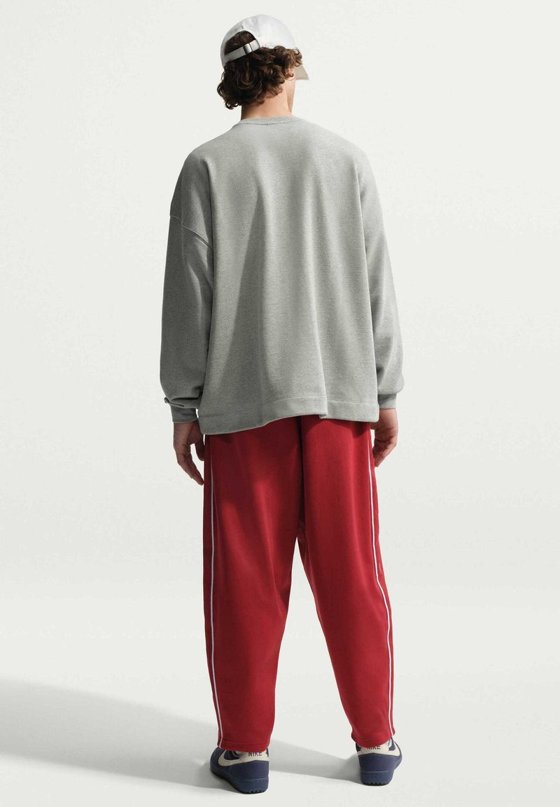 Personne portant une casquette blanche, un sweat-shirt gris et un pantalon rouge ample avec des rayures blanches, debout de dos sur un fond uni.