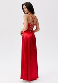 Červené saténové večerní šaty s dlouhým designem, které mají šněrování na zádech a tenké nastavitelné ramínka. Hladká textura a splývavá silueta.