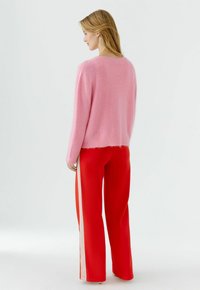 Frau mit blondem Haar, die einen rosa flauschigen Pullover und rote weite Hosen mit weißen Seitenstreifen trägt, seitlich auf weißem Hintergrund stehend.