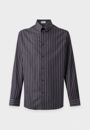 Un mannequin masculin se tient devant un fond uni et clair, vêtu d’une chemise noire à fines rayures, d’un jean bleu, d’une ceinture noire à boucle dorée et de mocassins noirs.