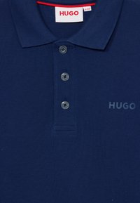 Polo bleu marine en tissu texturé avec une patte de boutonnage à trois boutons, un accent rouge à l'intérieur du col, et un logo "HUGO" imprimé.