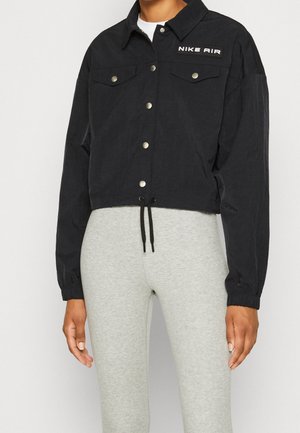 Mujer que lleva una chaqueta corta negra con botones de presión y el logo "Nike Air", combinada con leggings ajustados de color gris claro.