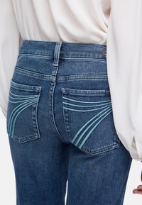 Achteraanzicht van een persoon die een blauwe spijkerbroek draagt met gebogen turquoise stiksel op de achterzakken en een losse witte blouse met lange mouwen.