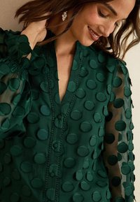 Zelená blůza s texturovaným designem a kruhovými látkovými akcenty, s předním zapínáním na knoflíky a průhlednými rukávy se sm gathered cuffs.